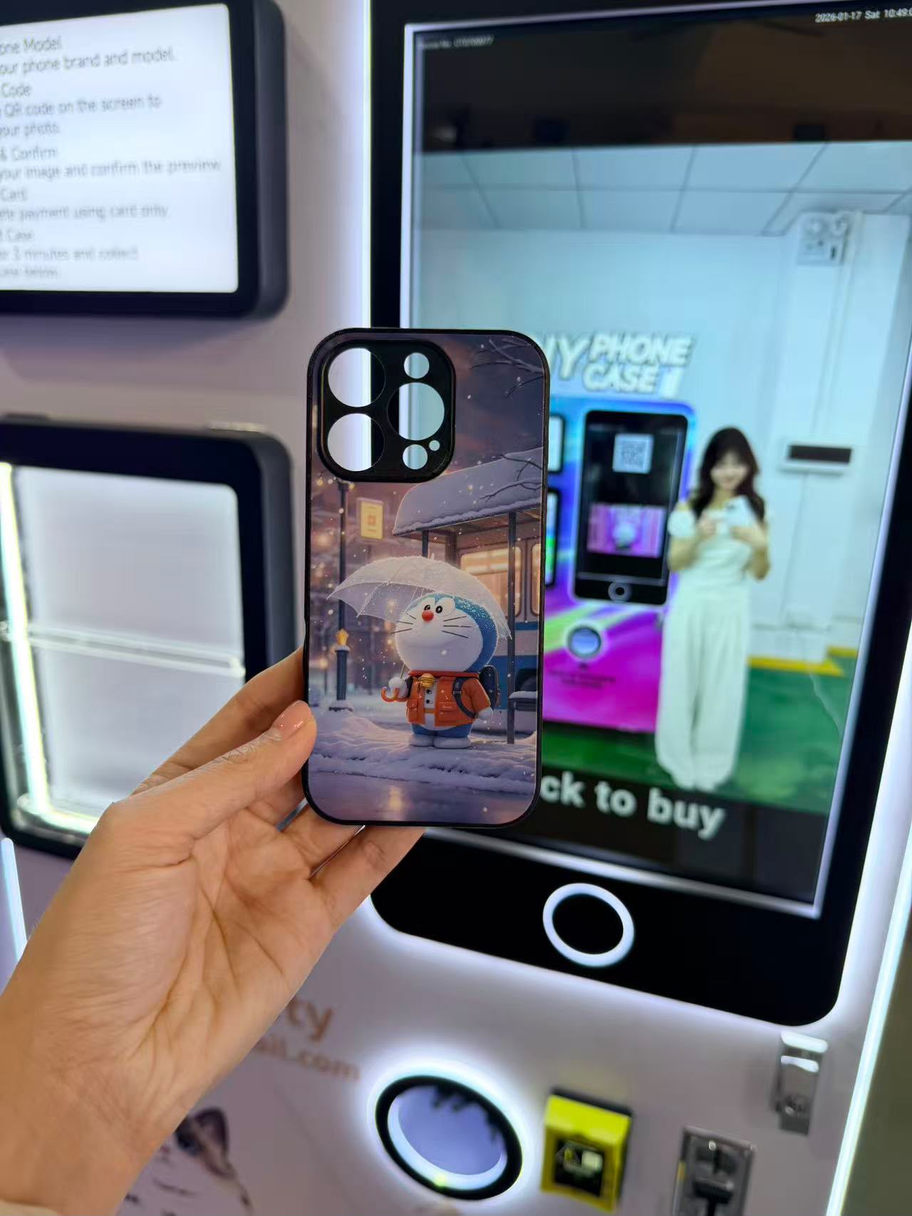 A customer holding a custom-printed phone case featuring a winter scene with Doraemon, produced by the DIY Phone Case vending machine in the background.